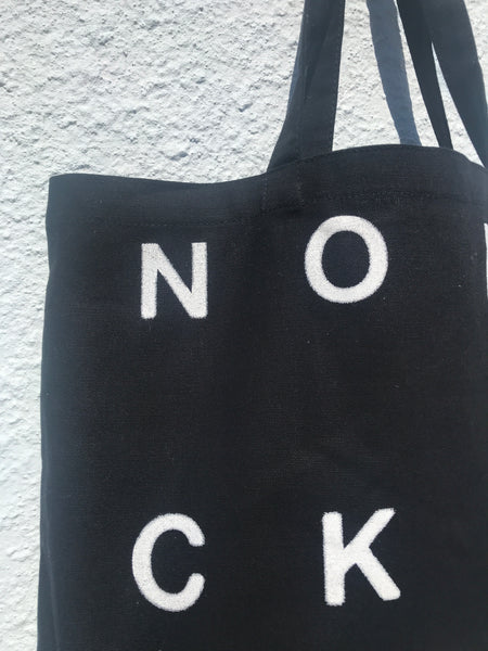 ZOOMED IN VIEW OF UPPER RIGHT CORNER OF BLACK TOTE BAG. PRINTED LETTERS N O C K IN WHITE ALSO VISIBLE.